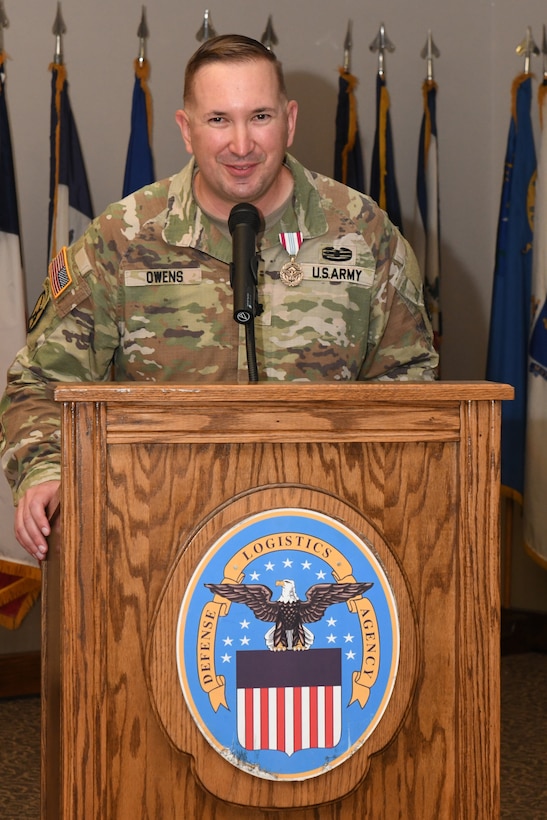 A man wearing a camouflage military uniform stands behind a lectern giving a speech.