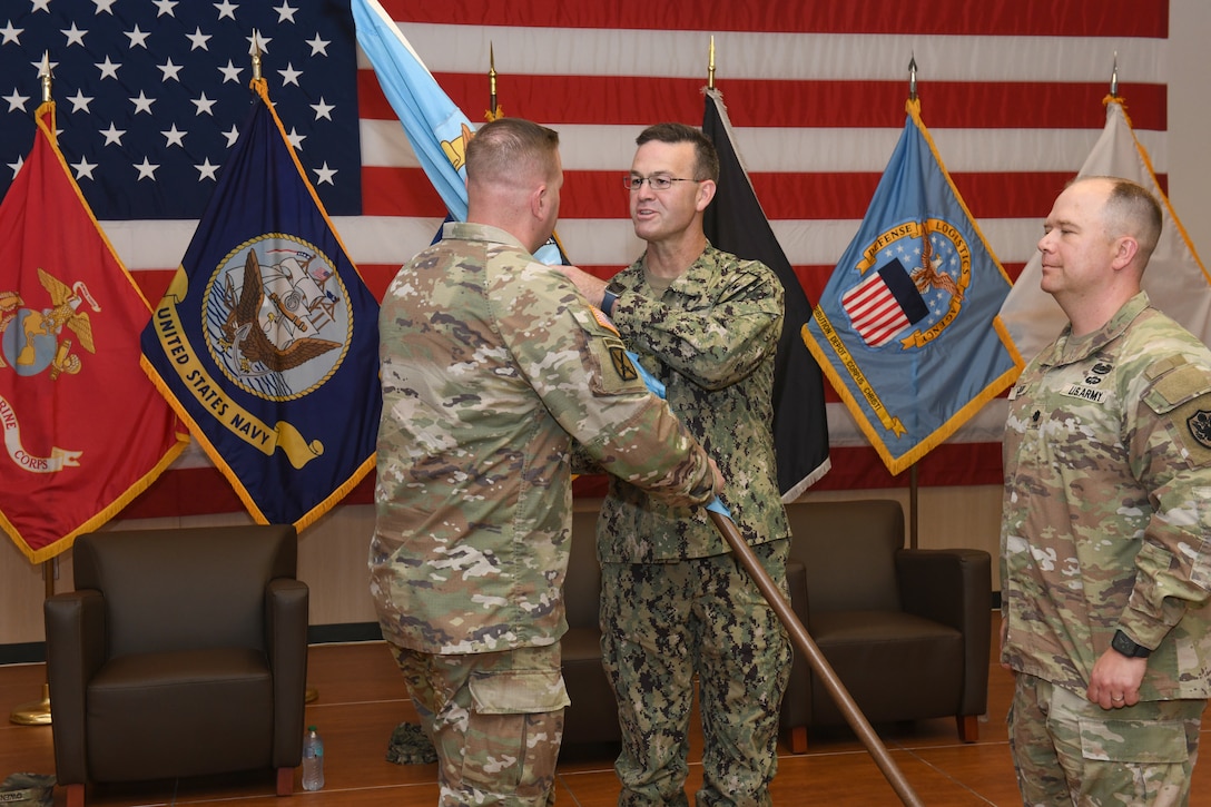 Three men wearing camouflage military uniforms stand on a stage with a United States flag and military branch flags behind them. Two of the men are passing a blue flag on a flagpole between them.