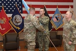 Three men wearing camouflage military uniforms stand on a stage with a United States flag and military branch flags behind them. Two of the men are passing a blue flag on a flagpole between them.
