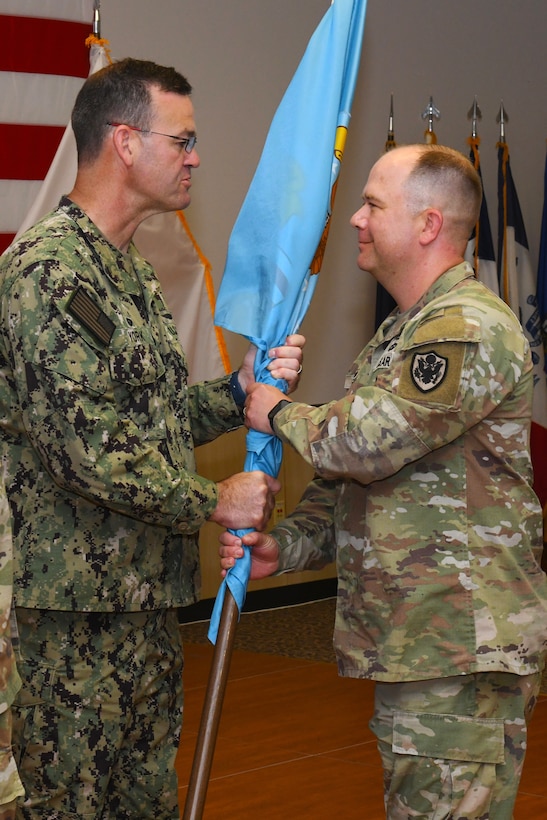 Two men wearing camouflage military uniforms pass a flag on a flagpole to each other.