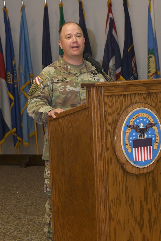 A man wearing a camouflage military uniform stands behind a lectern, speaking.