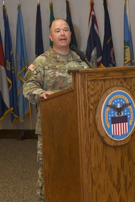 A man wearing a camouflage military uniform stands behind a lectern, speaking.