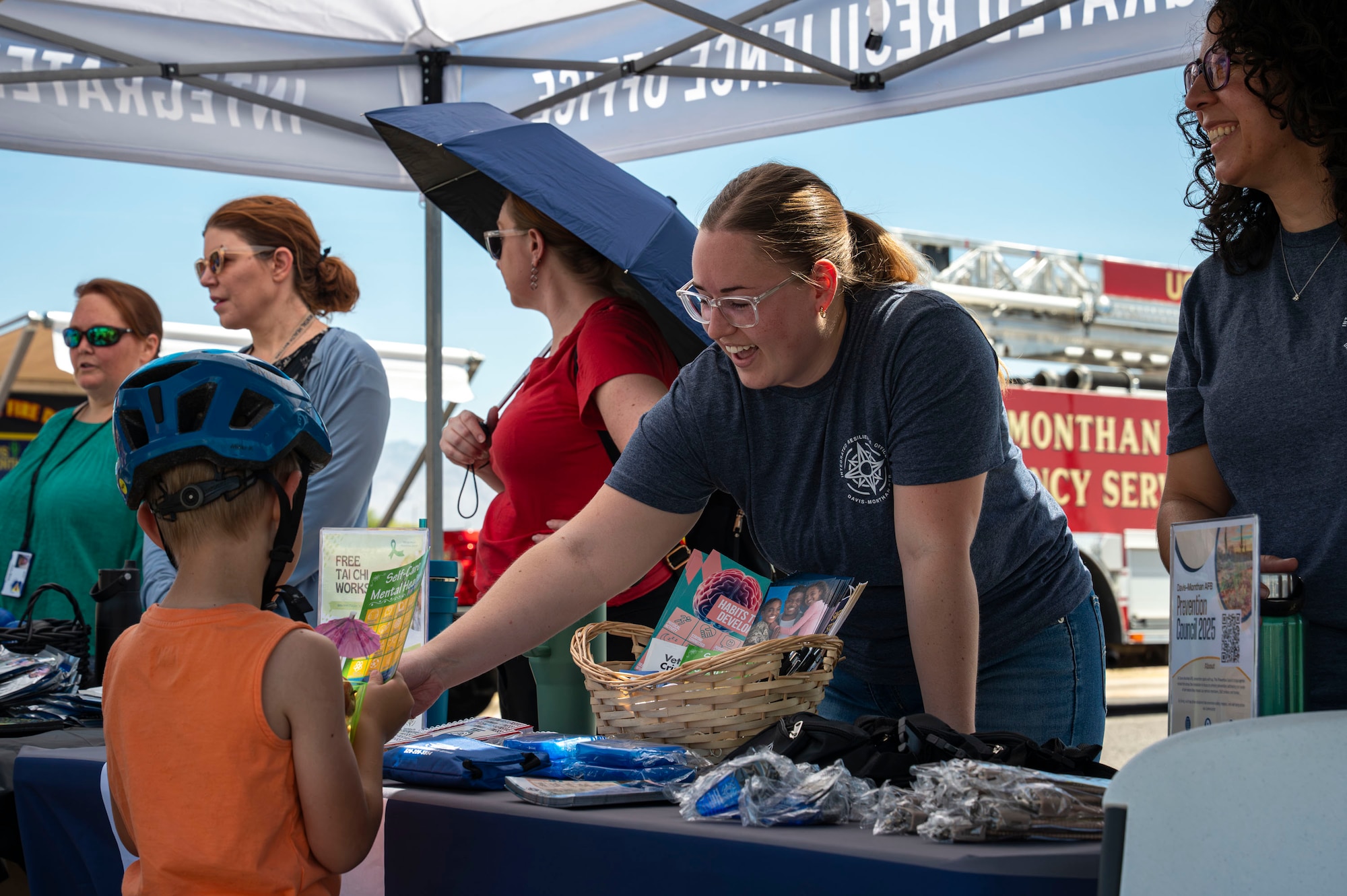 A resiliency booth volunteer gives a handout to a child during the Health Fair at Davis-Monthan Air Force Base, Arizona, May 28, 2025. The 355th Medical Group held the event to enhance operational readiness and promote physical, mental and spiritual health. (U.S. Air Force photo by Airman Samantha Melecio)