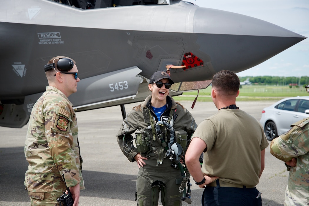 A female and two males in military uniforms talking with the nose of a plane in the background.