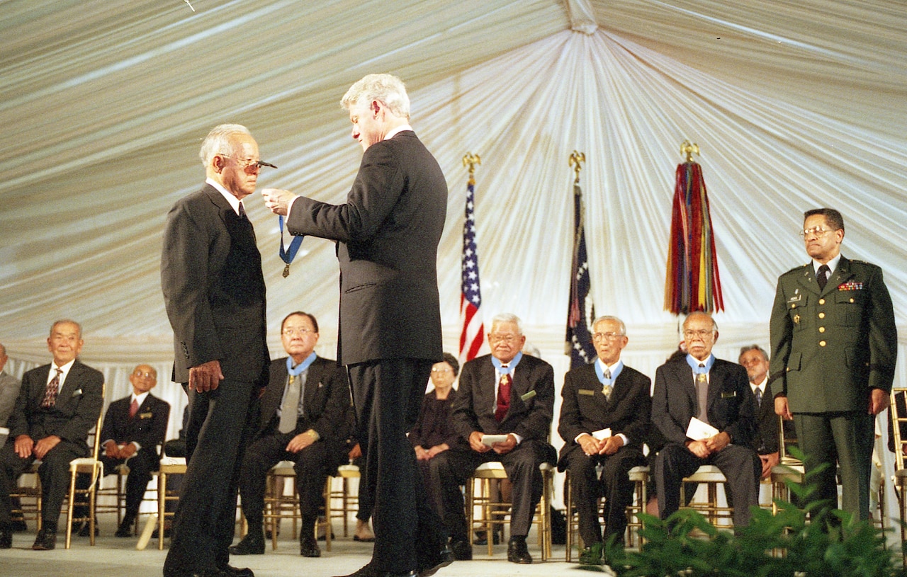 A man holds a medal on a ribbon in front of another man who is preparing to accept it around his neck. Others sit in the background, watching.