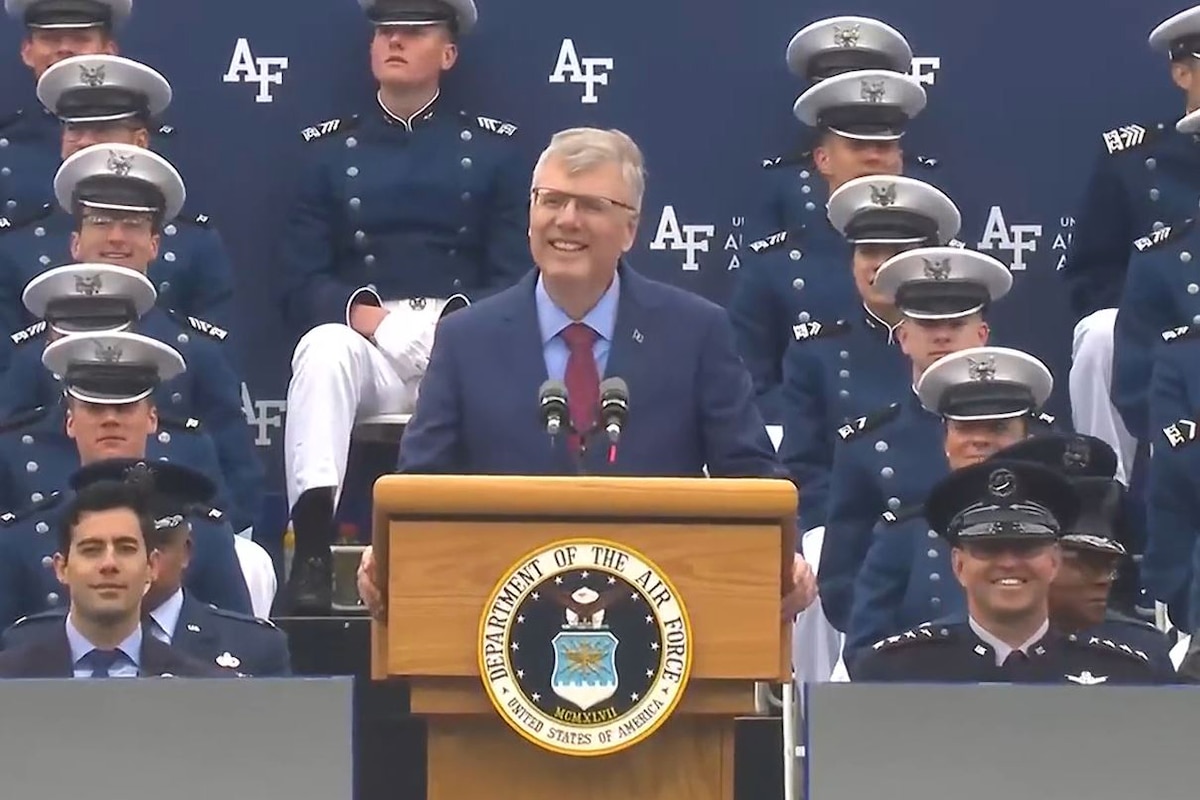 A man in a business suit speaks from a podium in front of military personnel in uniform.