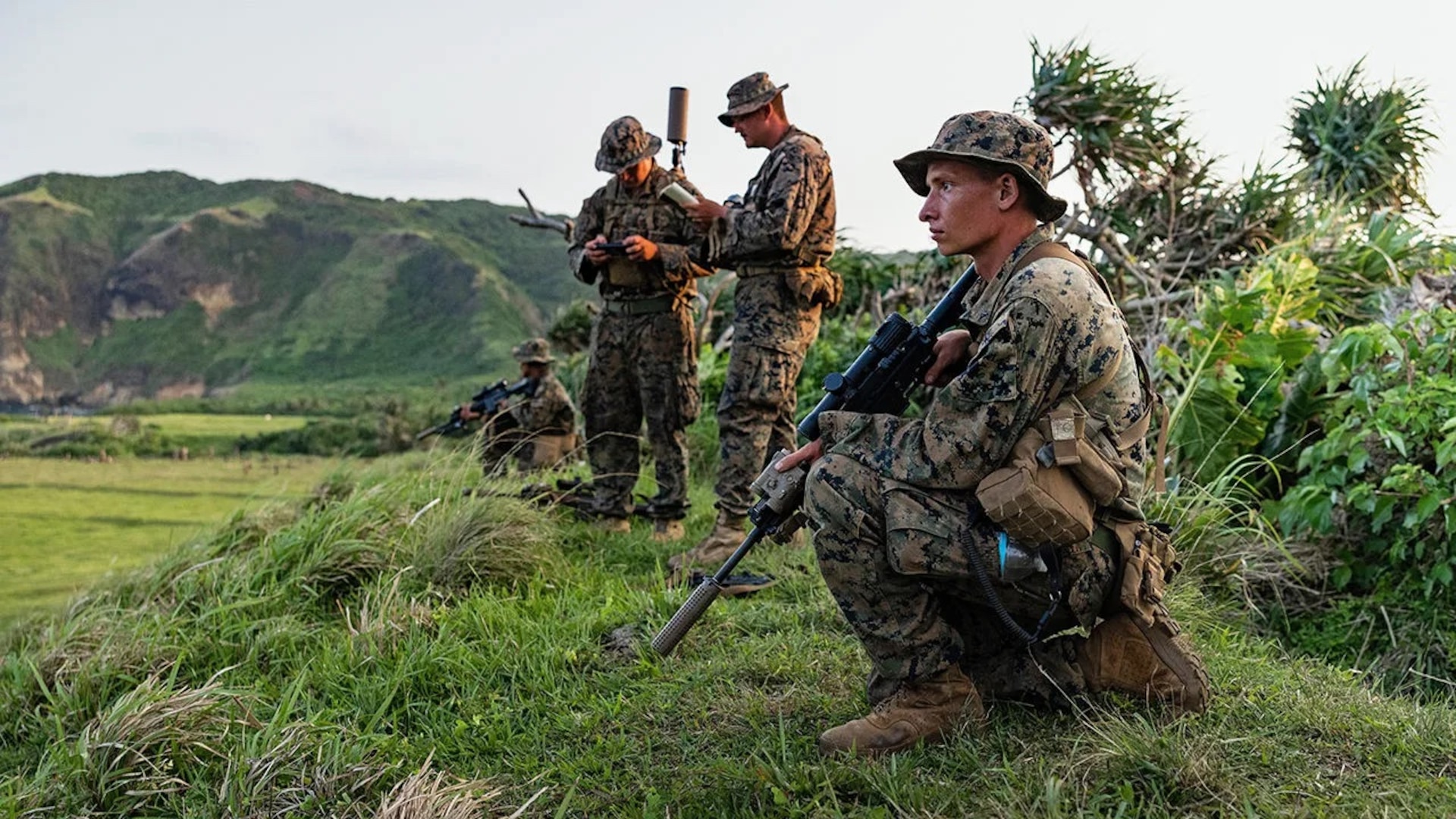 U.S. Marines in camouflage uniforms conduct field training on a grassy hillside with mountainous terrain in the background. One Marine kneels in the foreground holding a rifle with a scope, while others stand and work with communication equipment. The scene suggests a tactical or reconnaissance exercise in a remote, tropical environment.