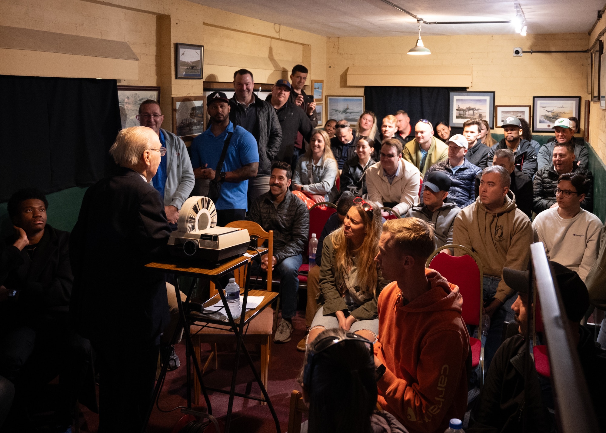 A crowd is gathered around an elderly man using a slide projector