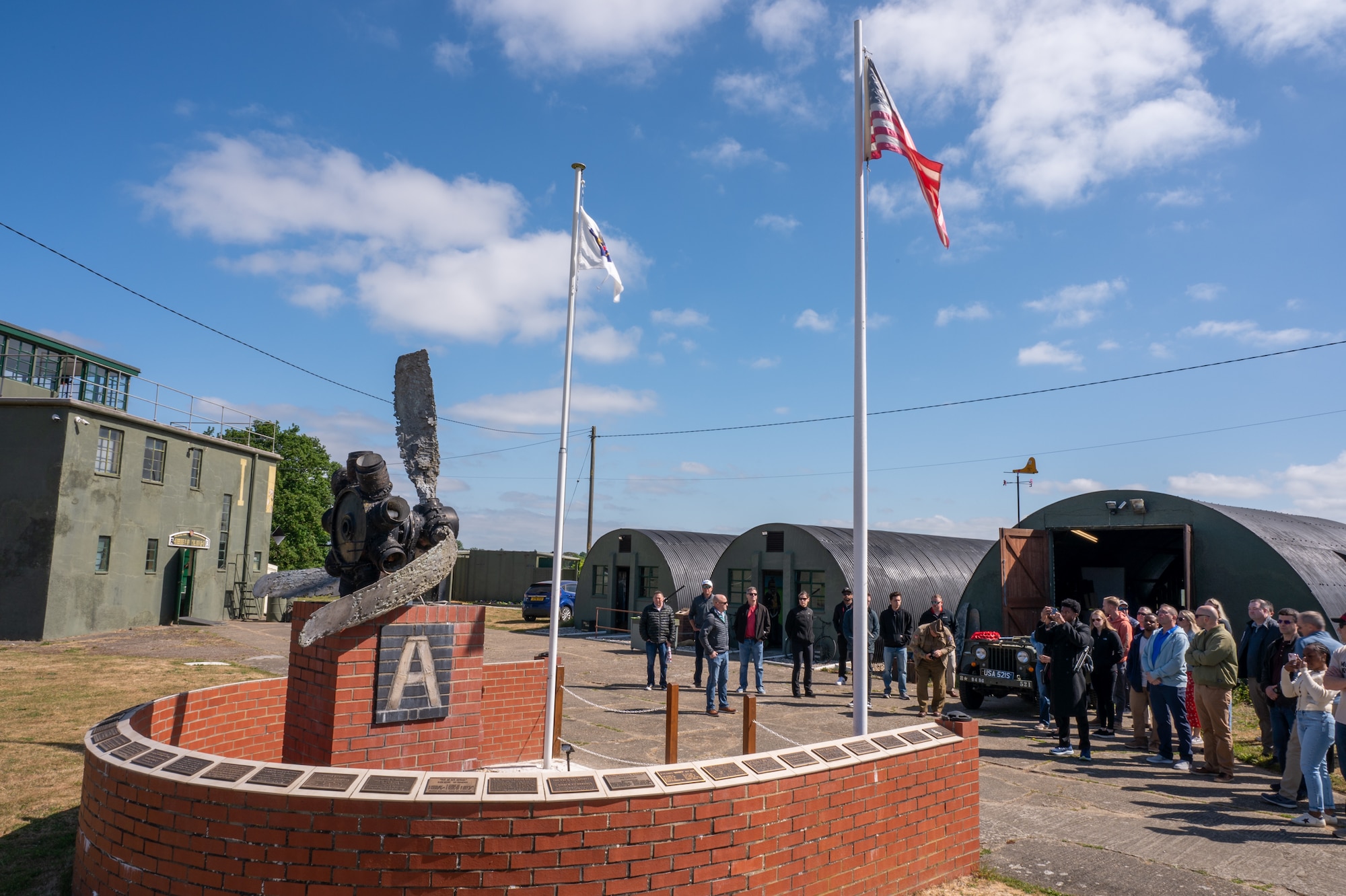 People stand near a memorial that features flags and a Big Sqaure A insignia at Rougham Control Tower Aviation Museum