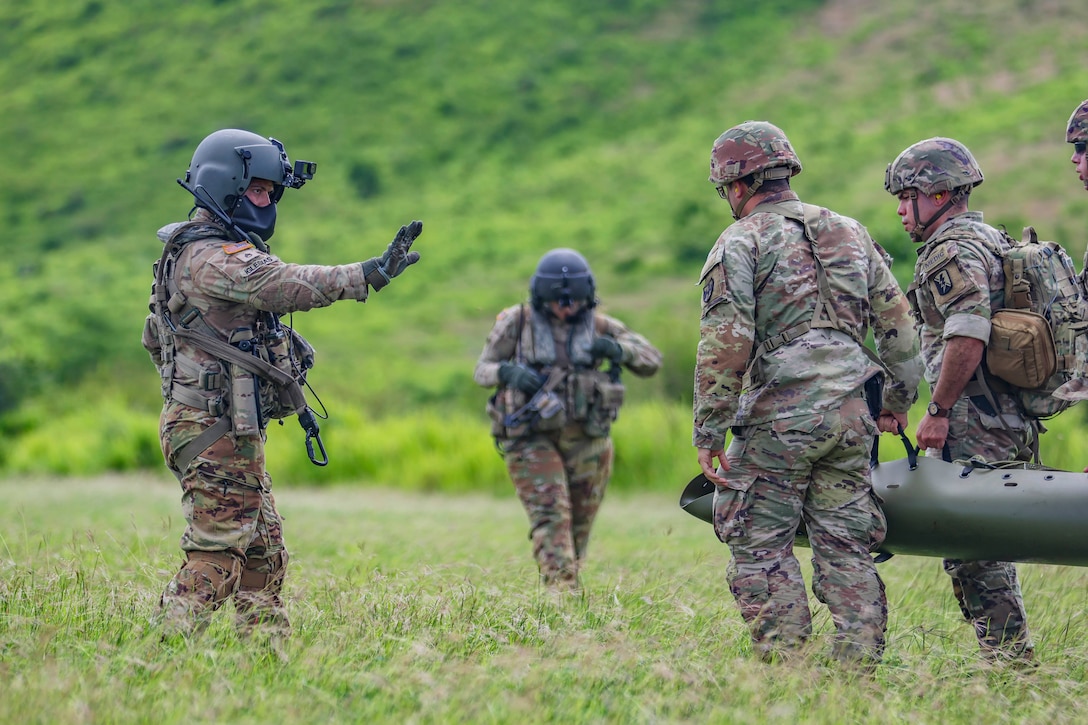 A soldier wearing protective gear leads three fellow soldiers carrying a dummy on a sling in a field.