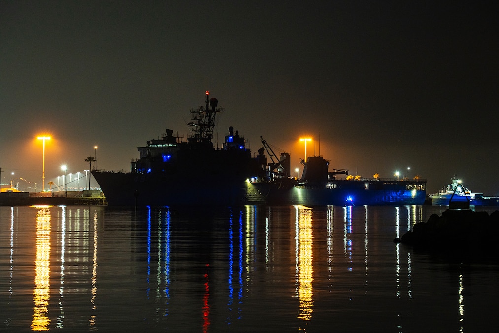 Ships and a boat are docked in the dark illuminated by orange and blue lights.
