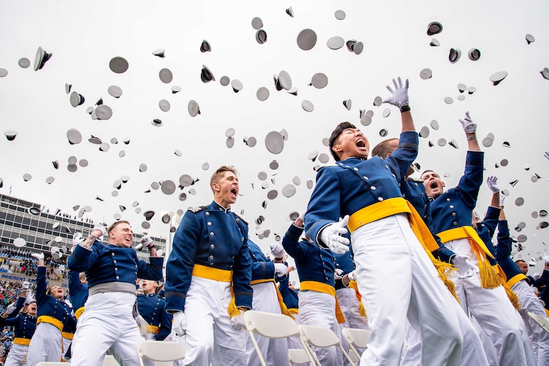 Hundreds of cadets in ceremonial dress cheer while tossing their caps at graduation at a stadium.