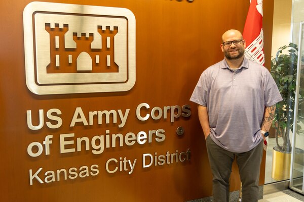 A man in a purple shirt stands in front of a brown wall with a silver emblem on the left side and a red flag on the right side.