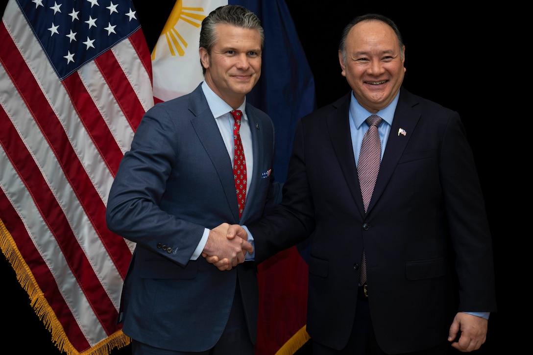 Two people wearing suits shake hands in front of the American and Philippine flags.