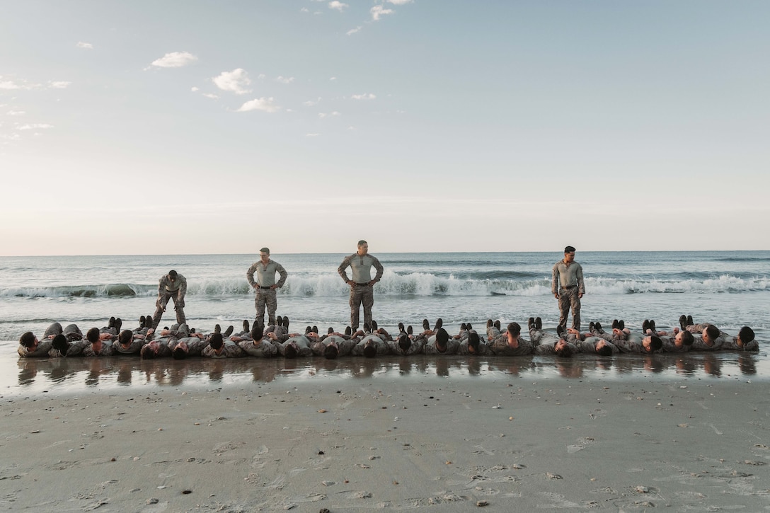 Four service members stand in front of a row of service members laying on a beach.