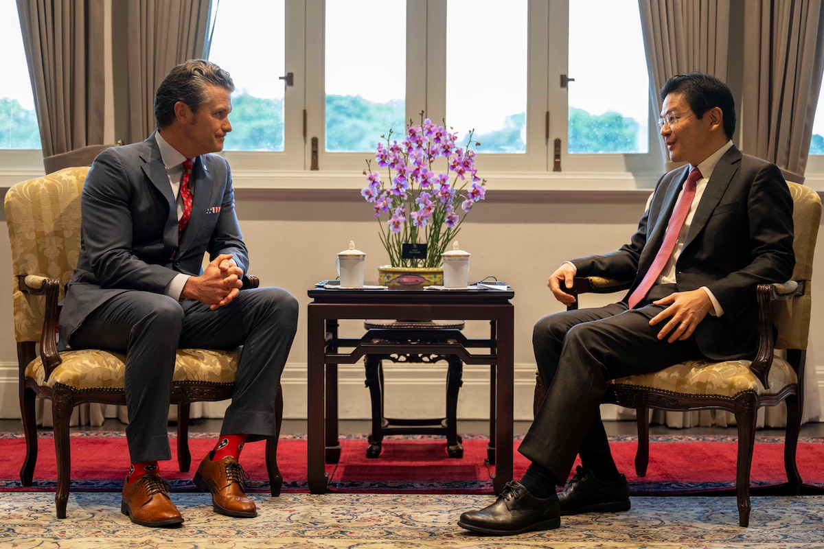 Two people wearing suits sit in front of windows and talk with a small table between them.