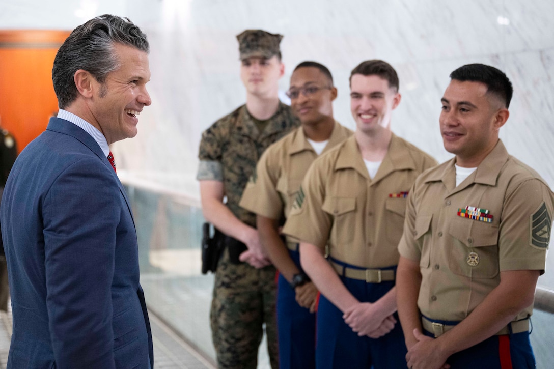 A person wearing a suit smiles at four Marines, three in dress uniforms and one in a camouflage uniform, standing indoors.