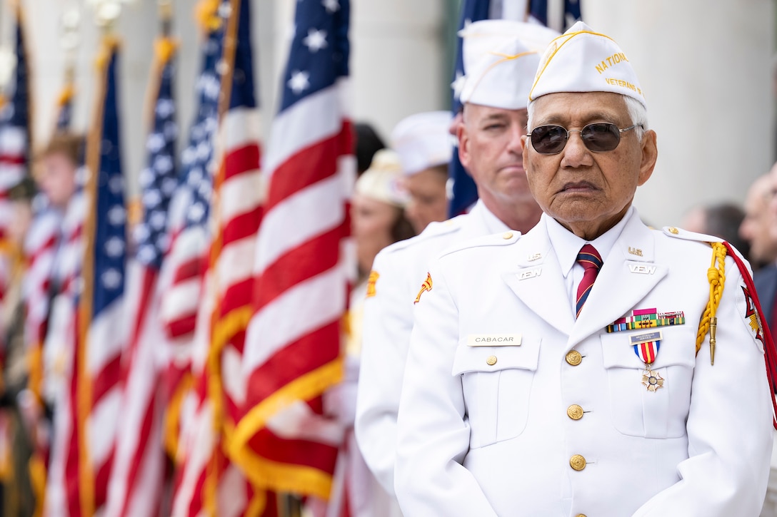 A veteran wearing a white uniform stands at the front of a procession with U.S. flags in the background outdoors.