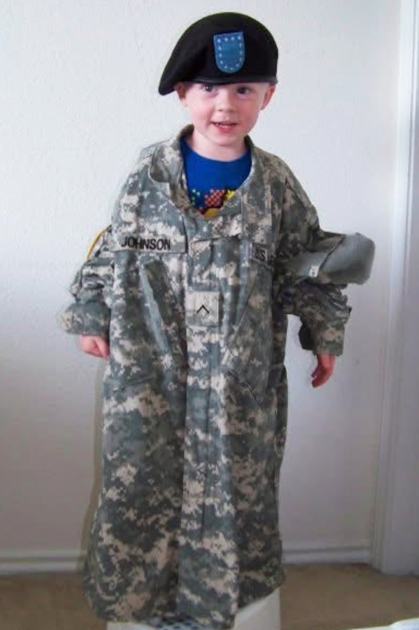 A toddler wears an adult combat shirt and beret.