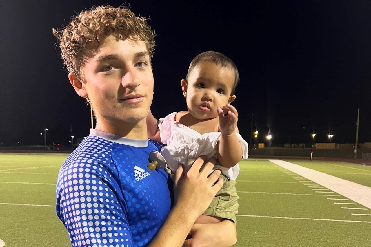 A boy in soccer uniform holds a baby on a field at night.