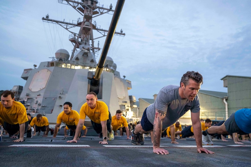 People wearing T-shirts and shorts perform pushups on the deck of a military ship.