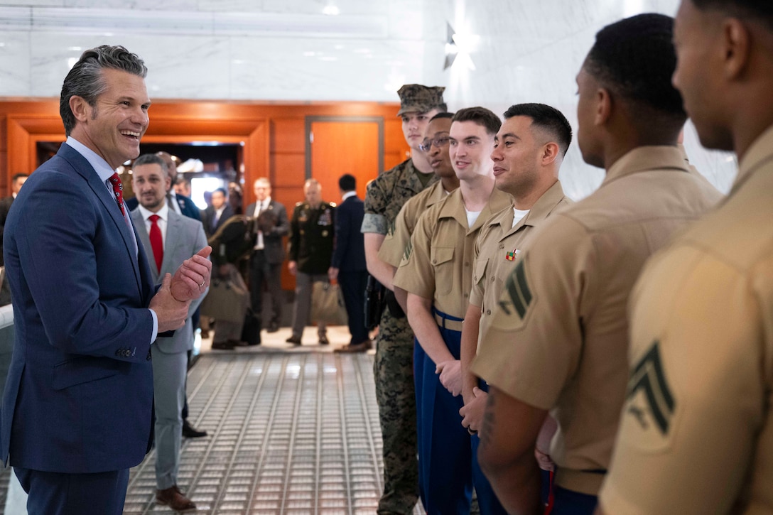 A person wearing a suit smiles at six Marines standing in a hallway as other people stand in the background.