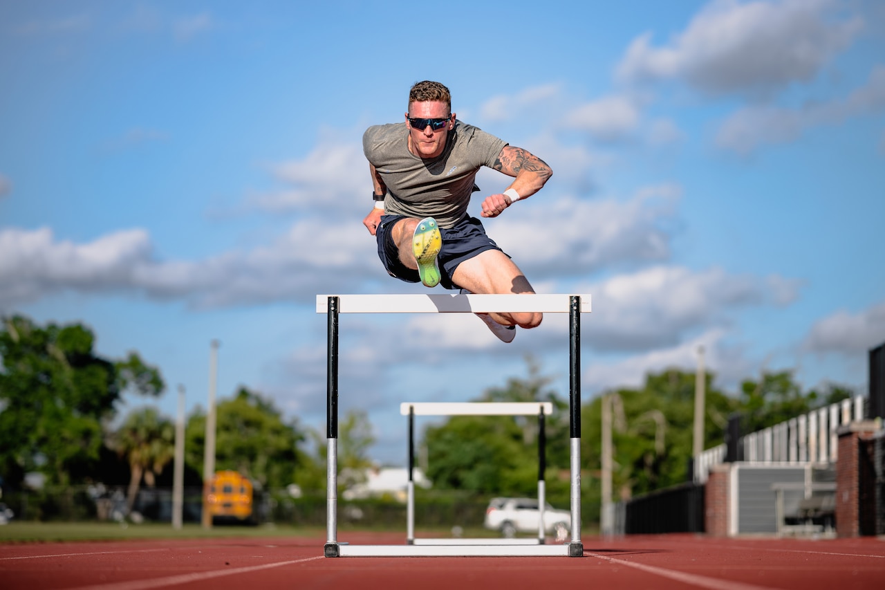 A decathlete jumps over a hurdle in a track field during a sunny day.