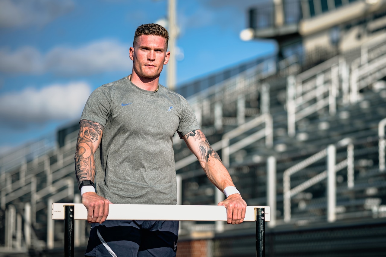 A decathlete poses for a photo holding a hurdle in front of stands.