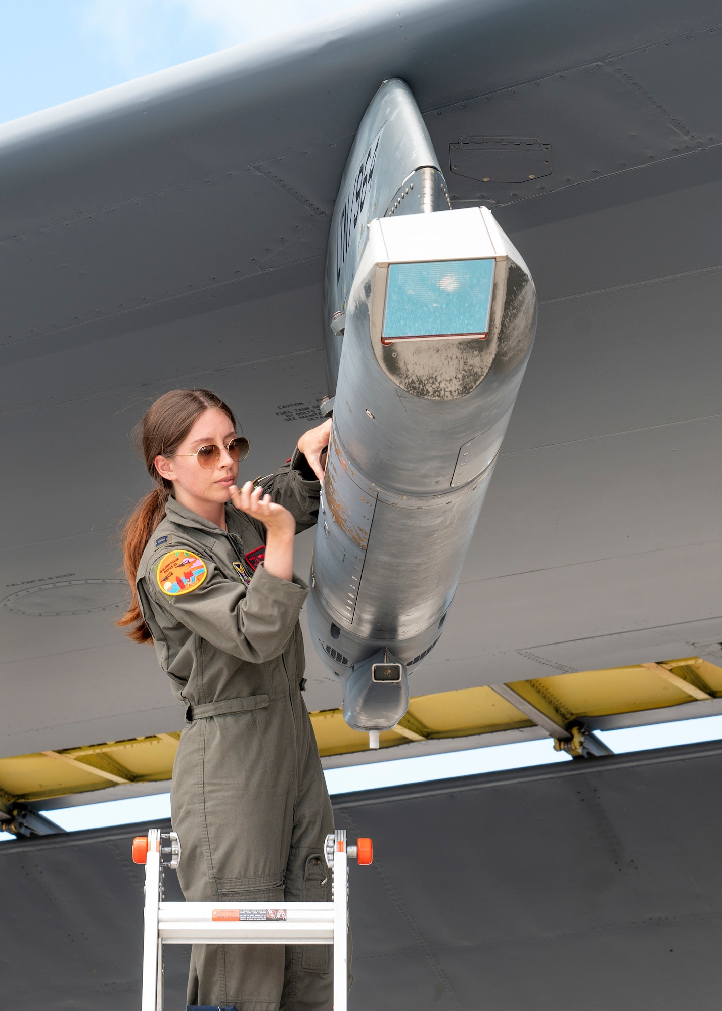 A female pilot wearing glasses adjusts a pylon under the wing of a very large military aircraft