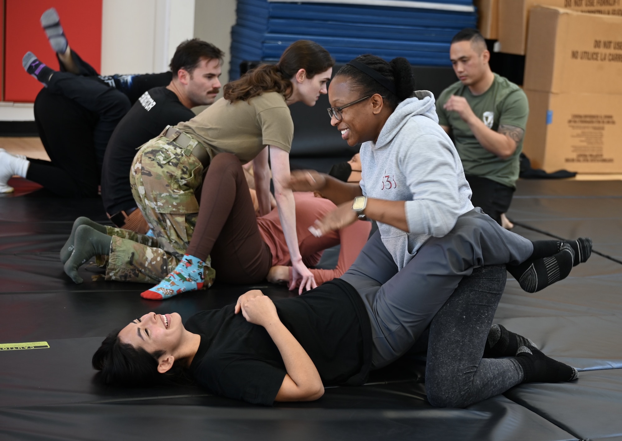 Several people in workout clothes are in pairs practicing defensive movement mechanics used in self-defense. One member of the pair lies on their back on a black mat with their legs wrapped around their partner, and the other member is on their knees.