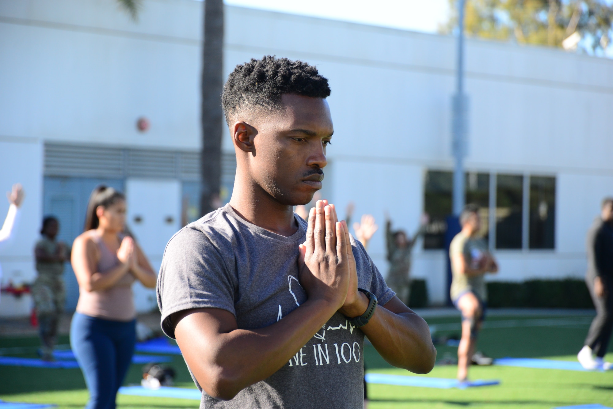 A man with black hair almost closes his eyes as he holds his hands, with his palms together while he is meditating. In the background, and out of focus, are people in the same pose standing on blue yoga mats on green grass.