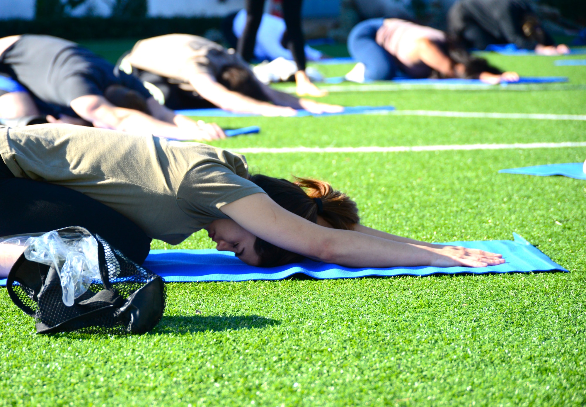 Four people on blue yoga mats stretch in a resting yoga pose called child's pose. They are on green grass of a soccer field on Los Angeles Air Force Base.