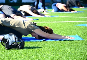 Four people on blue yoga mats stretch in a resting yoga pose called child's pose. They are on green grass of a soccer field on Los Angeles Air Force Base.