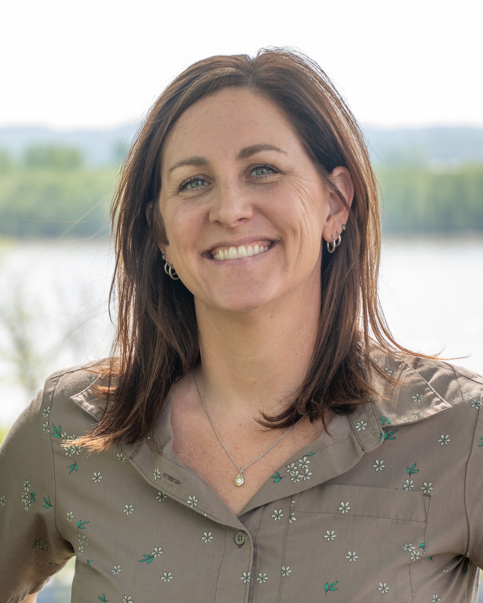 A smiling woman poses for a photo in front of the Mississippi River. She is wearing a brown blouse.