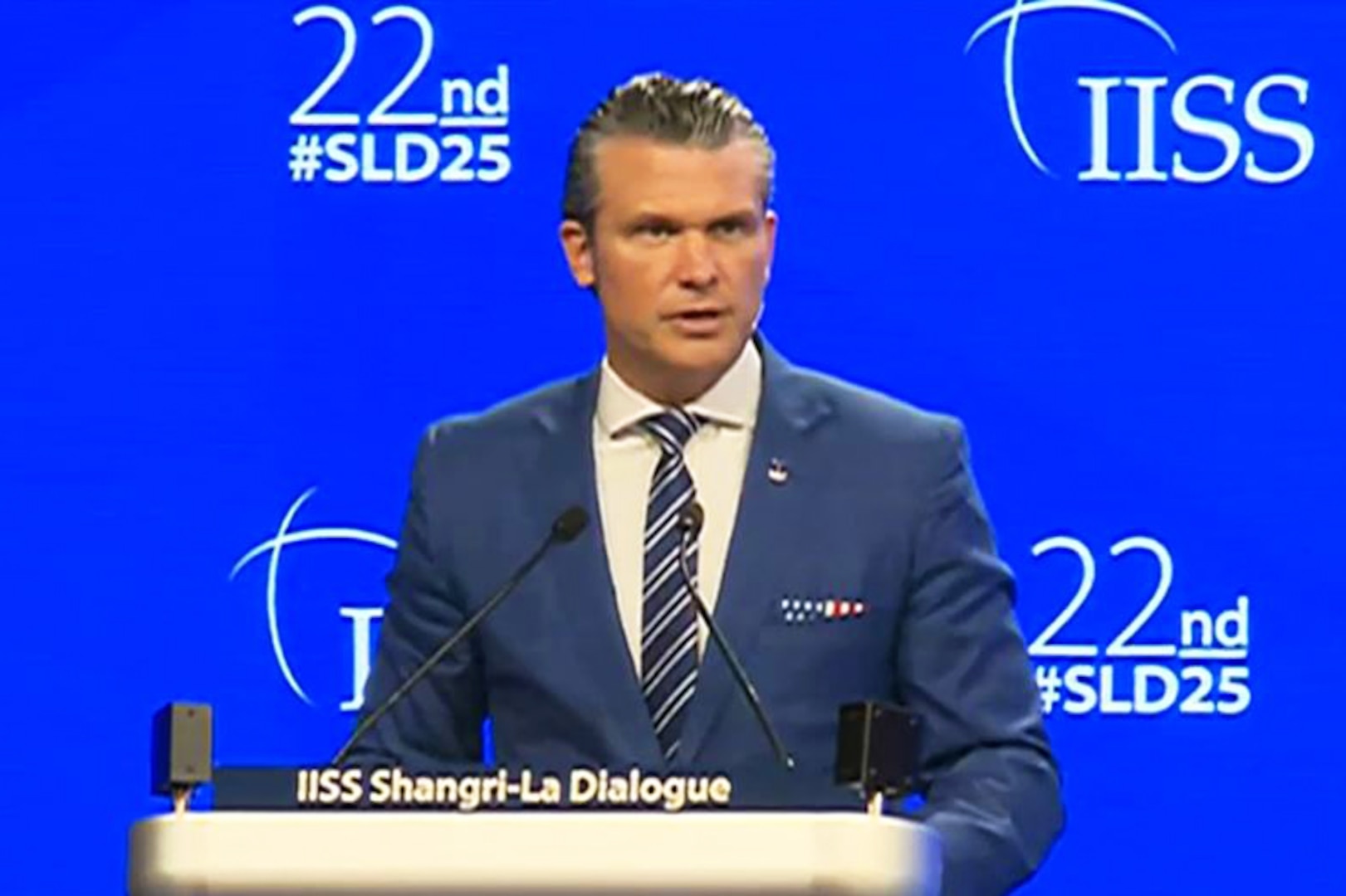 Defense Secretary Pete Hegseth speaks into two mics at a lectern with a bright blue background and the white text: ISS 22nd #SLD25