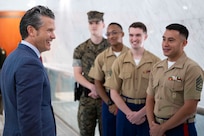 A person wearing a suit smiles at four Marines, three in dress uniforms and one in a camouflage uniform, standing indoors.