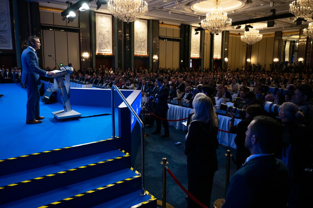 A man in a suit stands at a lectern on a stage in front of an audience in a lecture hall.