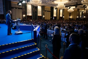 A man in a suit stands at a lectern on a stage in front of an audience in a lecture hall.