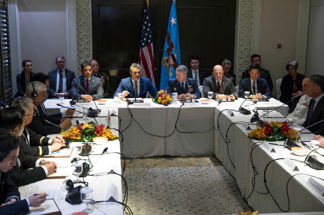 A group of men in suits sit around a rectangular table in a room with two flags.