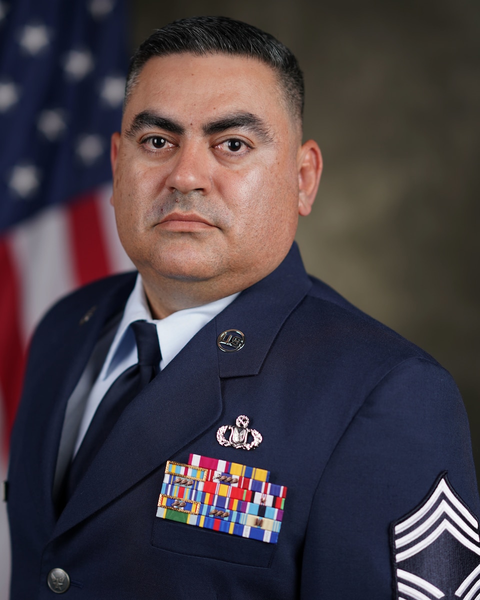 An official Air Force biography photo of the interim Command Chief, a head and shoulders portrait from the chest up, depicting Chief Master Sgt. Enrique Lopez, facing the camera at a 45 degree angle depicting an official Air Force background drop behind him with a U.S. flag  draped behind his right shoulder.