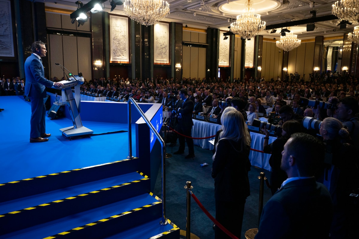 A man in a suit stands at a lectern on a stage in front of an audience in a lecture hall.