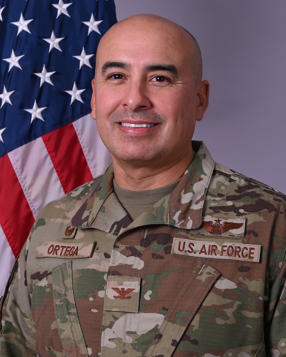 A man in a camouflage military uniform smiles for a photo in front of an American flag.