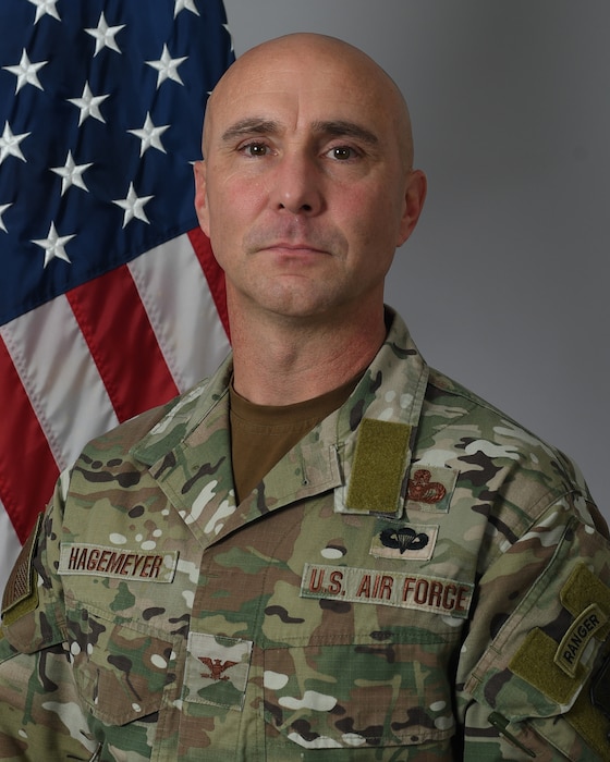 A man in a camouflage military uniform stands in front of an American flag.