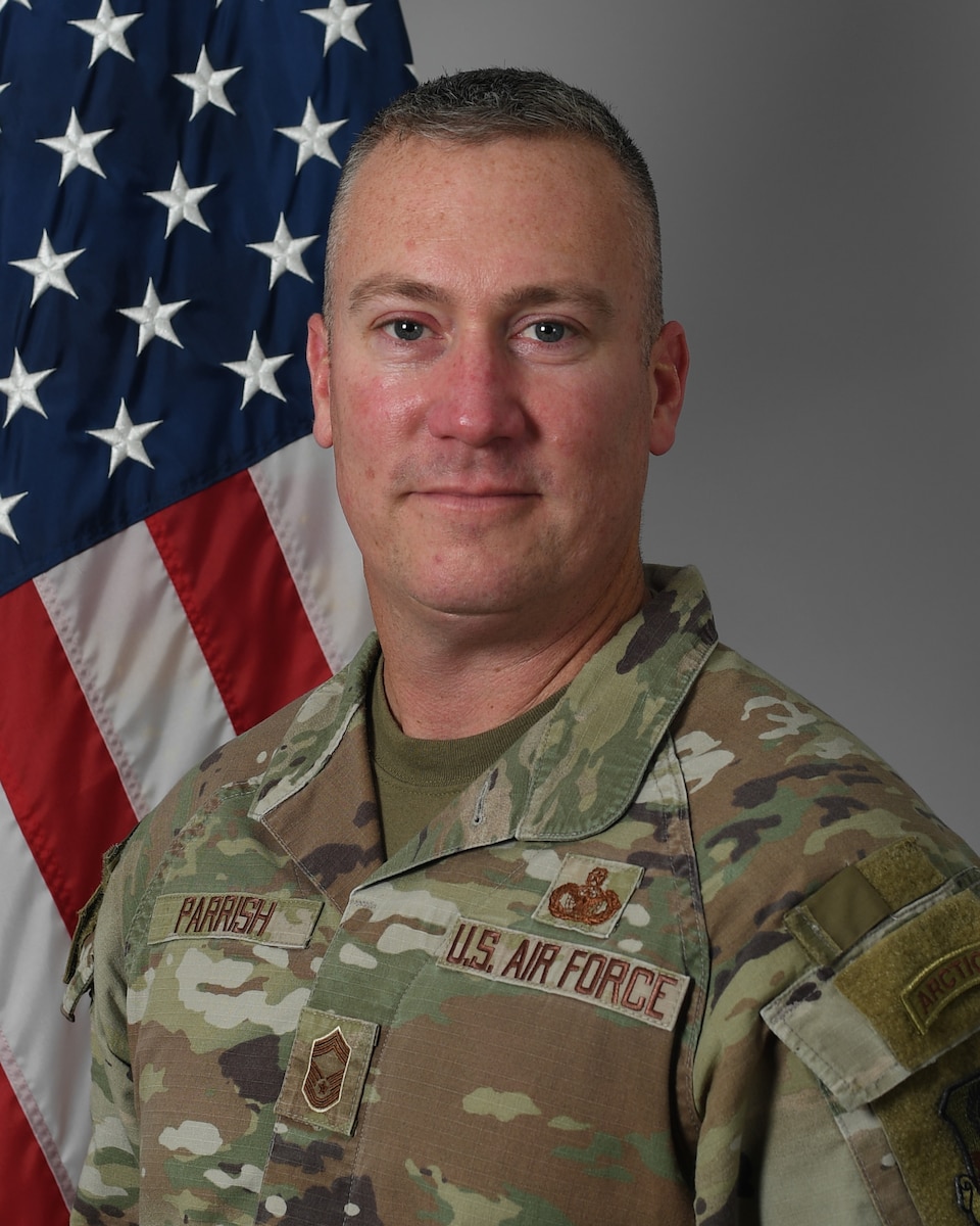 A man in a camouflage military unform stands in front of an American flag.