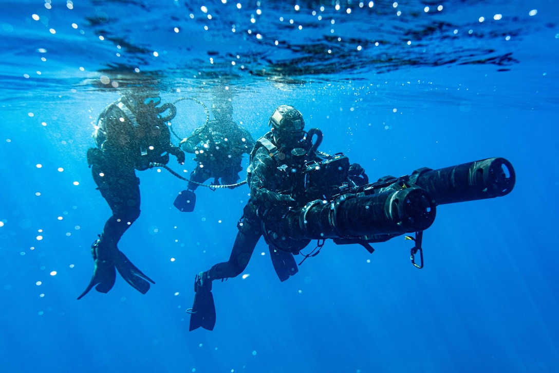 Three sailors in dive gear swim in blue water, one holding a long black device.