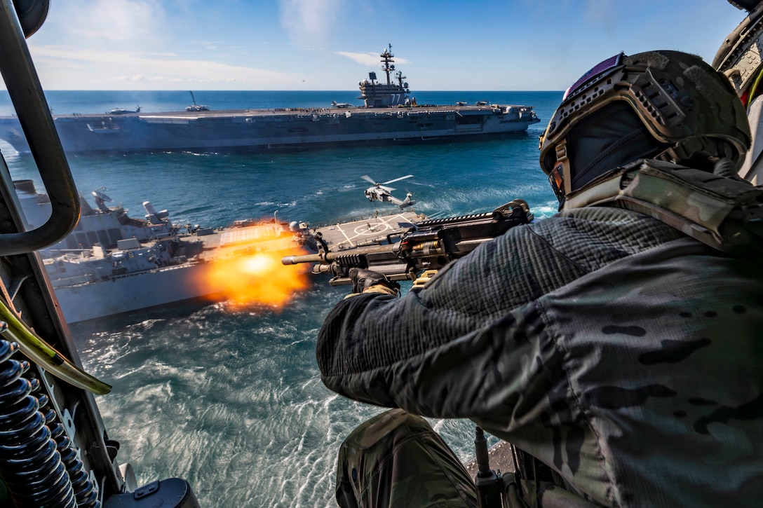 A sailor shown from behind fires a weapon from the open door of a helicopter flying near two ships at sea.