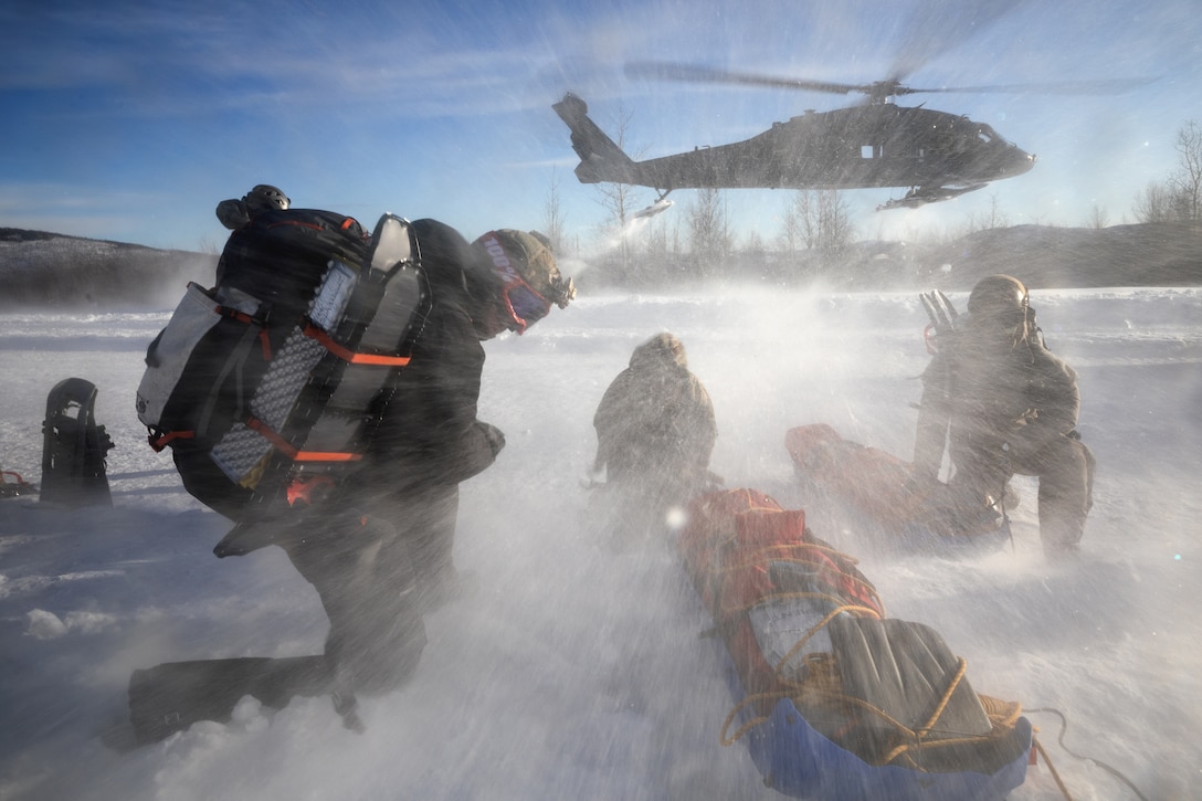 Service members in winter gear kneel on a snowfield as snow sprays them from a helicopter hovering nearby.