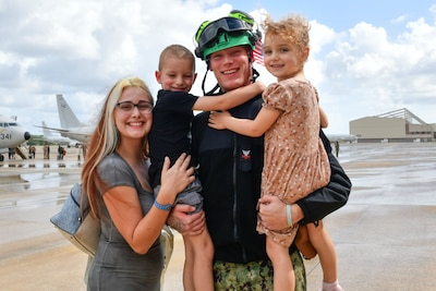 Uniformed service member stands with wife and two children in his arms in front of airfield