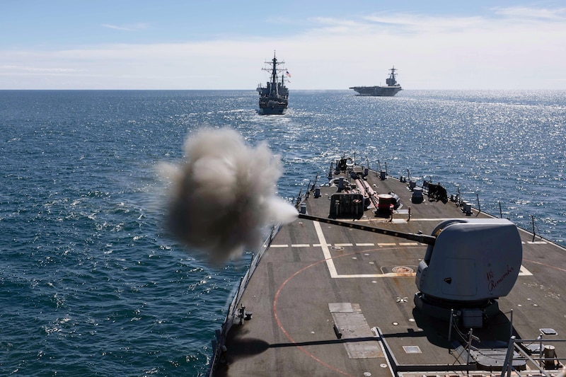 ISmoke billows from a machine gun on the deck of a ship with an aircraft carrier and another ship in the foreground.