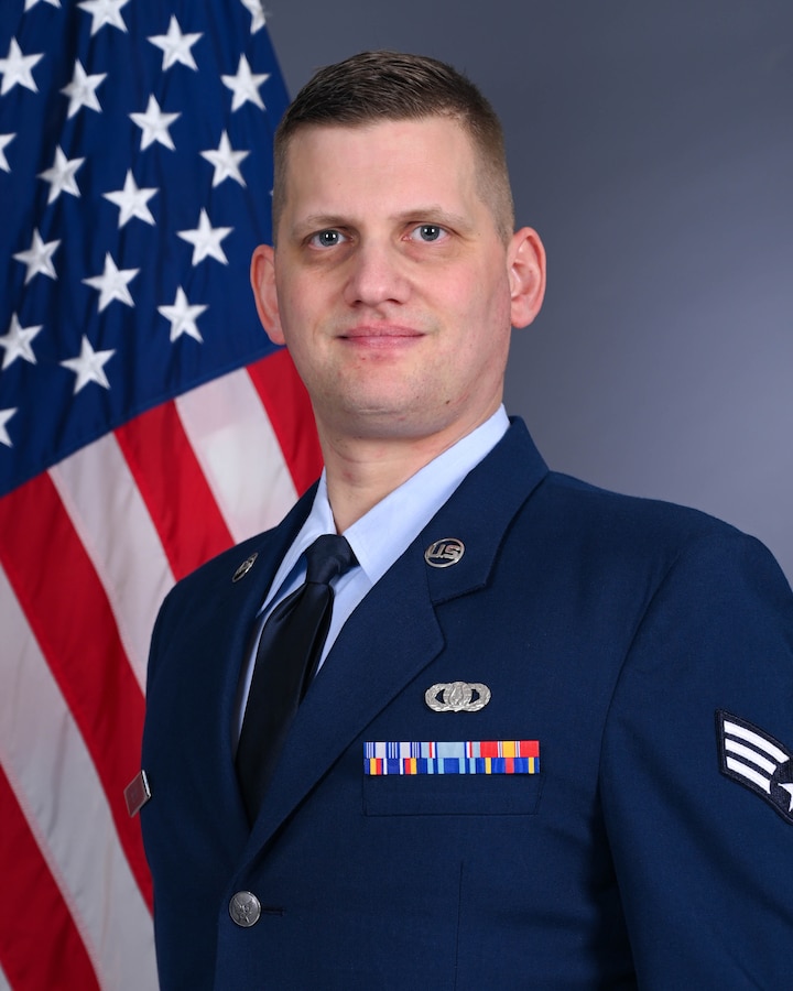 Official photo of Senior Airman Sebastian Bell for unit website wearing the service dress uniform standing in front of the American flag.